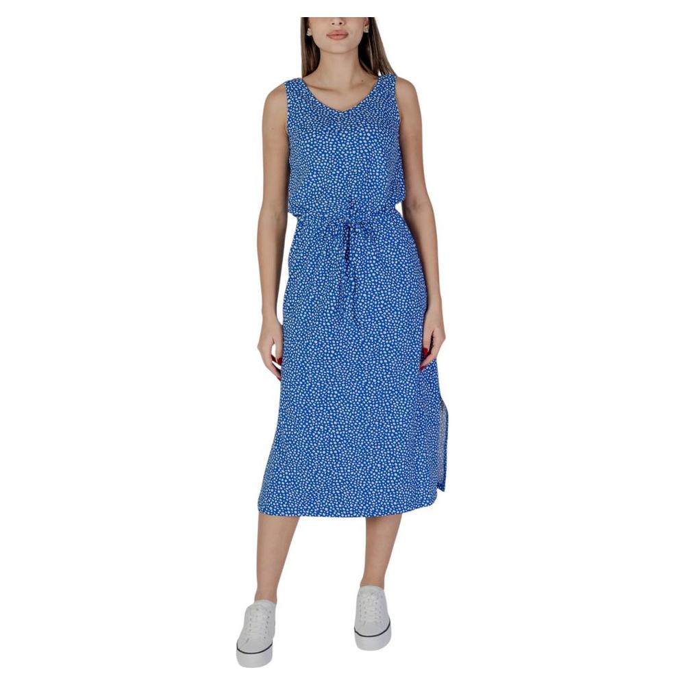 Woman wearing a sleeveless blue polka-dot dress with a waist tie, paired with white sneakers, standing against a plain background.