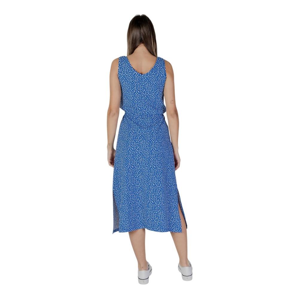 Back view of woman in blue sleeveless dress with white polka dots, mid-length, and side slits, paired with white shoes.
