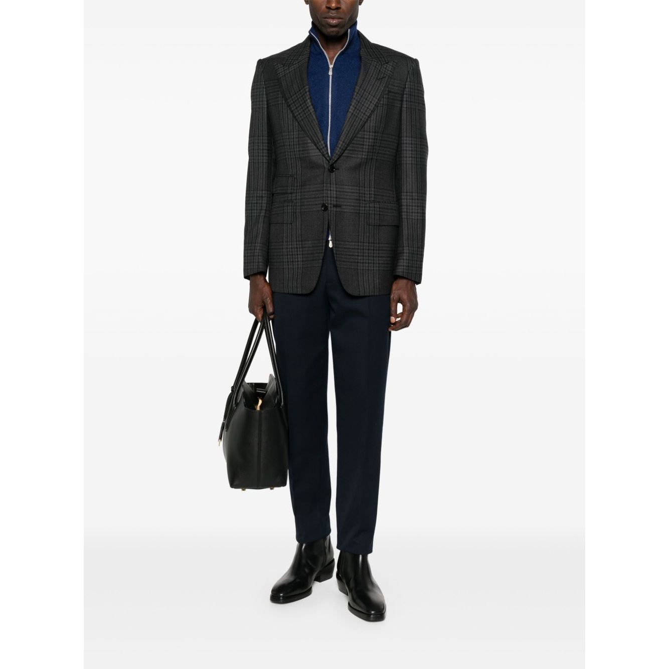 Model wearing a blue Brunello Cucinelli Cashmere zip-up sweatshirt under a gray blazer, paired with black trousers and holding a bag.
